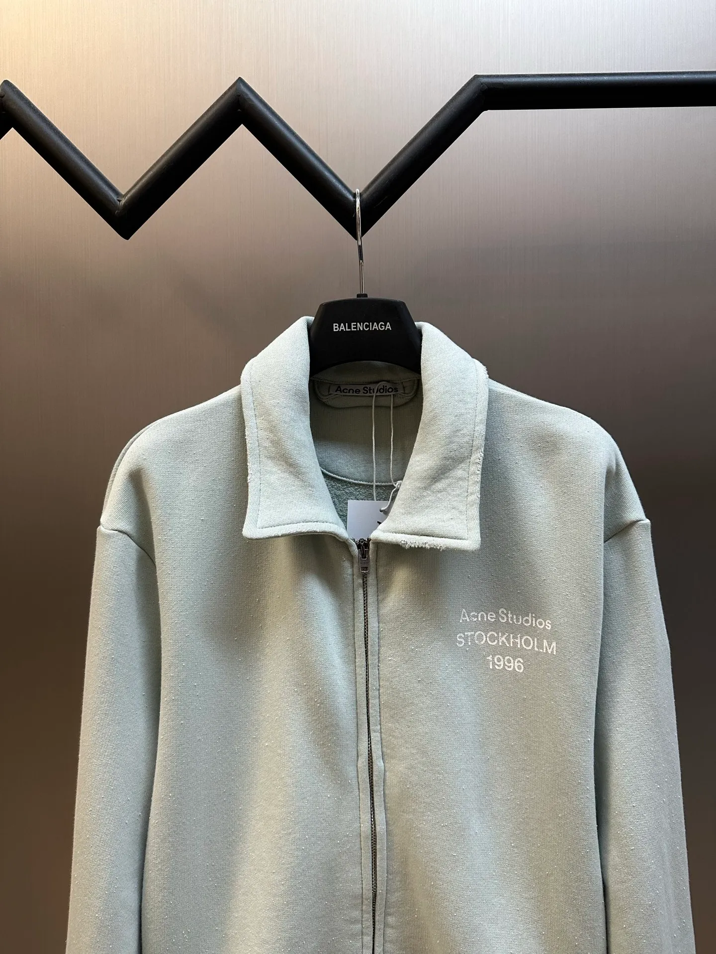 Ac vintage collared zipper sweatshirt uses 32-count top layer and 40-count bottom layer of 3-strand composite fleece structure, with a weight of g/㎡. The 32-count yarn on the top layer is fine and smooth, giving the surface a smooth touch and excellent anti-pilling properties; the 40-count 3-ply yarn on the bottom layer is treated with a raising process to form a rich and soft fluff layer, providing excellent warmth and skin-friendly wrapping. With 21 2×2 ribs, the g/㎡ weight ensures that the collar, cuffs and hem have excellent tensile fatigue resistance and shape retention capabilities. During piece dyeing, the entire fabric is immersed in the dye vat after weaving, and the dye molecules fully penetrate into the fibers, ensuring full, deep colors and excellent color fastness. Each batch of dyeing produces subtle differences, making the color tone of the single product unique. On this basis, the paint is sprayed irregularly on the surface of the garment through manual spray painting to create a splash-like retro effect. The distressed processing of holes causes the color to fade and mottle in parts. The ribbed trim is also treated to form slight whitening and fluffing, which visually echoes the spray-painted effect on the body, making the new clothes appear as if they have been worn for a long time and exposed to the sun. The front logo stamp uses a slurry printing process. The printing slurry contains a special microcapsule foaming agent. After being baked at high temperature, the slurry layer expands to form three-dimensional bulges, and the surface presents a granular texture similar to inkjet paint. This process simulates the effect of random drops of pigment after being atomized during spray gun spraying. The edges of the pattern present a natural feathering transition, rather than the rigid boundaries of glue printing. The printing surface has a fine graininess and soft elasticity to the touch, forming a closed loop of the process with the distressed effect of the spray painting on the body. size：xs s m l
